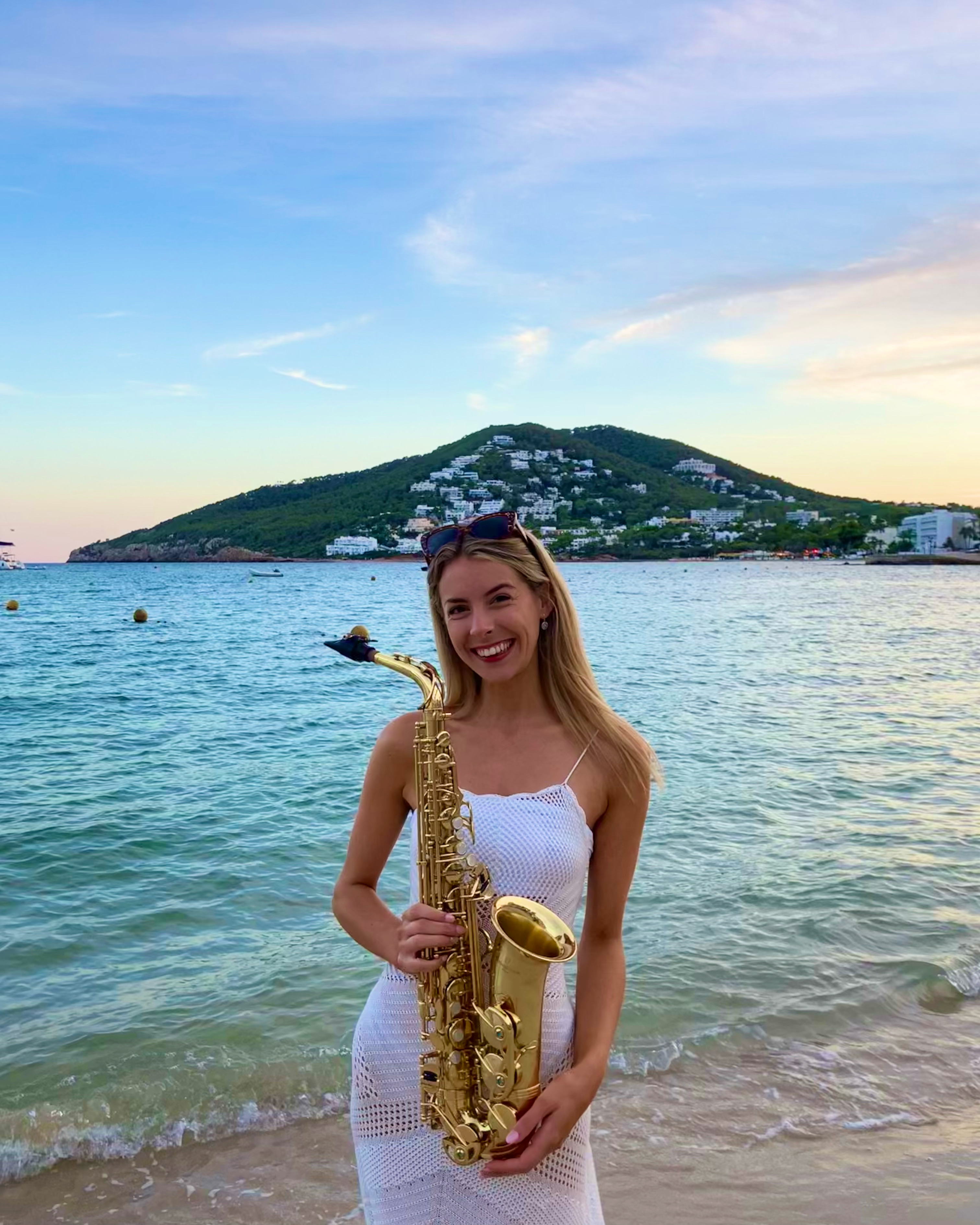 Blonde woman in white dress smiling and holding a saxophone in front of the turquoise Mediterranean Sea.