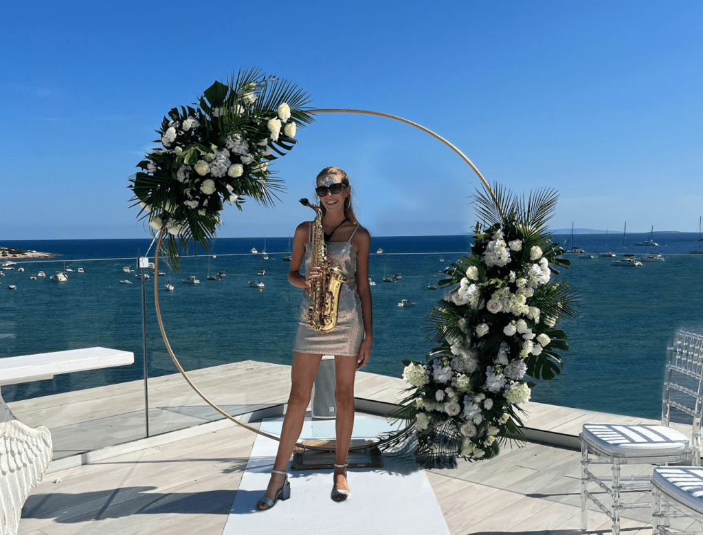 Smiling blonde woman with sunglasses in short, sparkly dress holding saxophone. She's standing on a rooftop in front of a circular flower decoration, with blue sea and sky in the background.