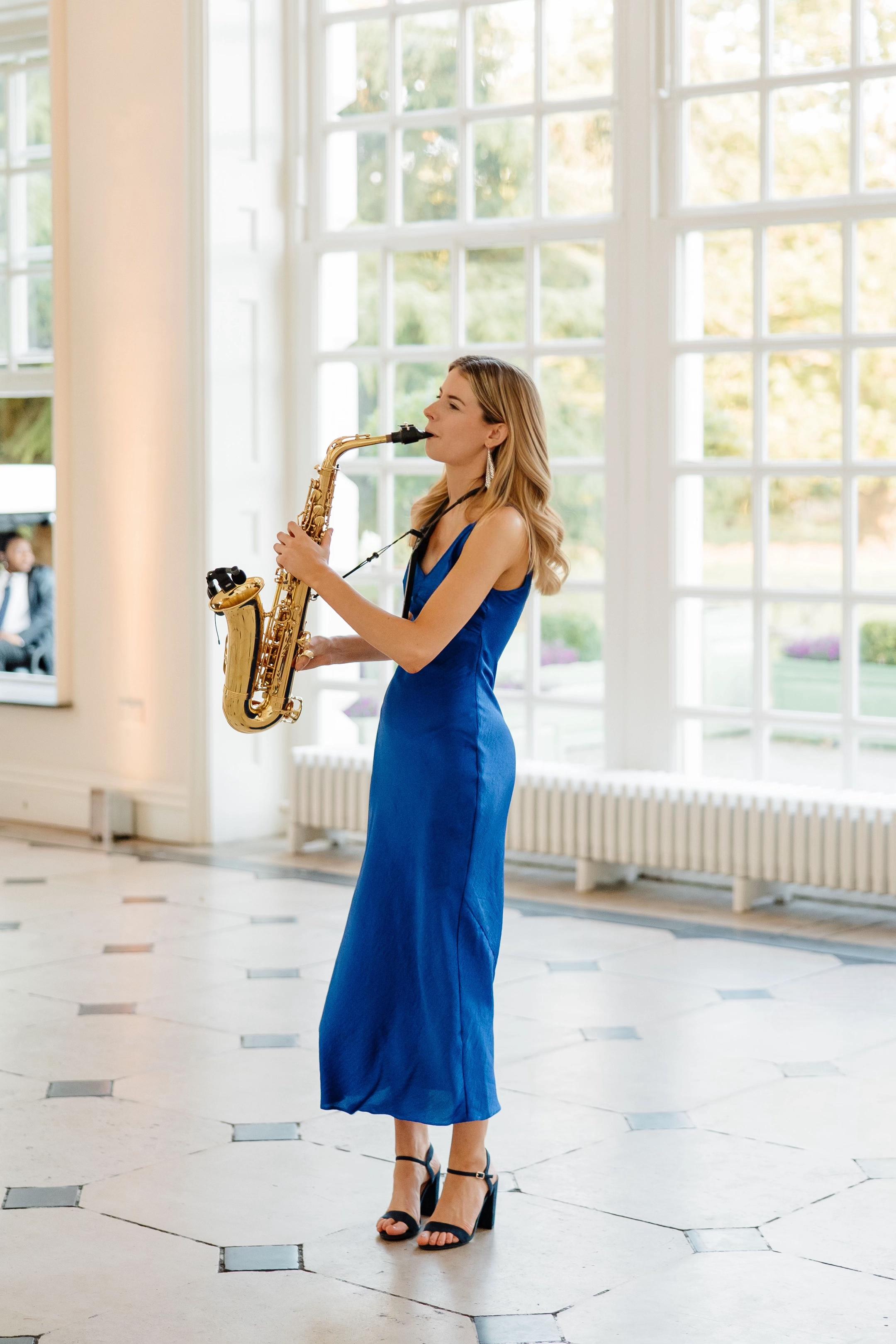 Blonde haired woman in long blue dress plays saxophone in a bright hall in front of a big window.